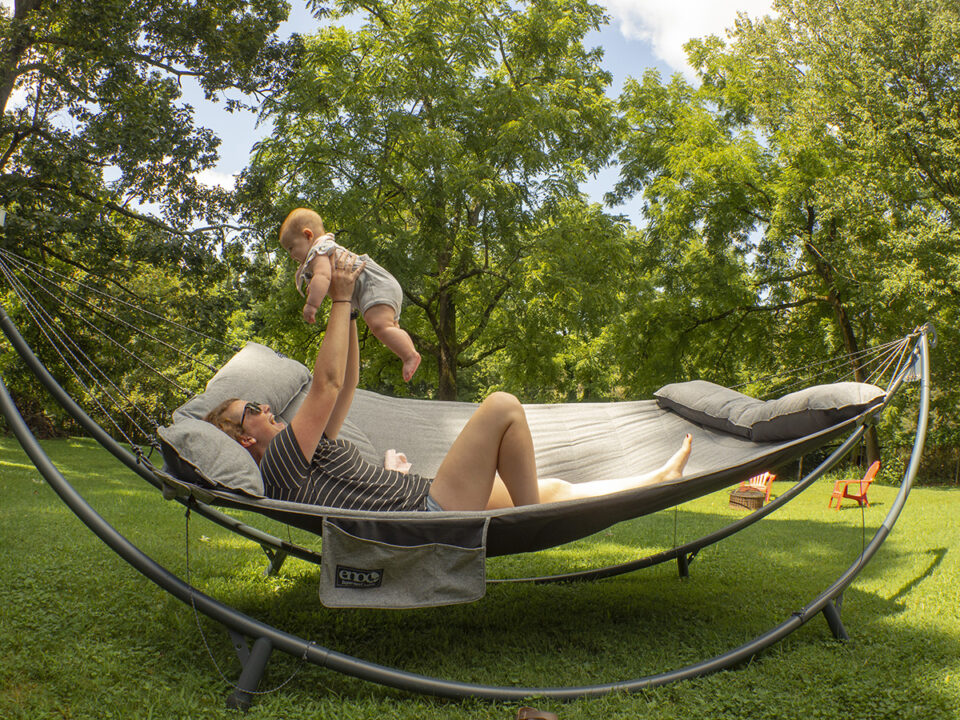 A woman raises her baby into the air as she lays on the SuperNest Hammock in the SoloPod XL Hammock Stand in her backyard.